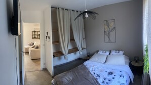 Un dormitorio con una cama, una mesita de noche, una lámpara colgante, una planta en la pared y una puerta que da a otra habitación.
