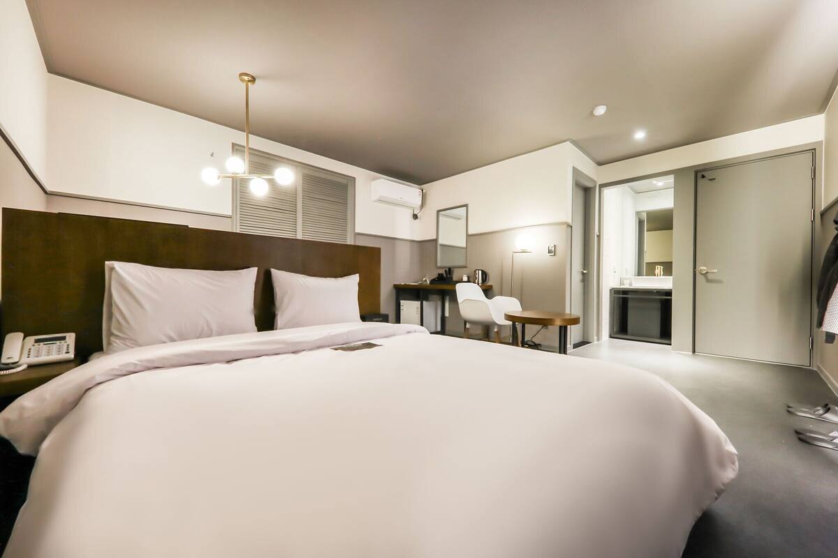 A modern hotel room with a large bed, a desk, a chair, a mirror, and a wardrobe.