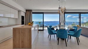 Une cuisine moderne avec un îlot central en bois, un coin repas avec des chaises bleues et de grandes fenêtres offrant une vue sur la mer.