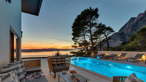 Un espace aménagé au bord de la piscine, avec des chaises longues, une table où est servi un dessert, et une vue sur la mer et les montagnes.
