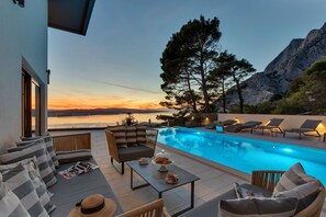Area piscina con poltrone relax, un tavolo con un dessert e vista sul mare e sulle montagne.