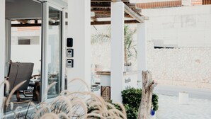 Uma área de estar moderna ao ar livre, com entrada por porta de vidro, pérgola de madeira e um muro de pedra ao fundo.