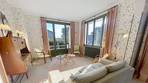 En stue med sofa, lænestole, et bord og et skab. Store vinduer med gardiner, en radiator og en lampe.