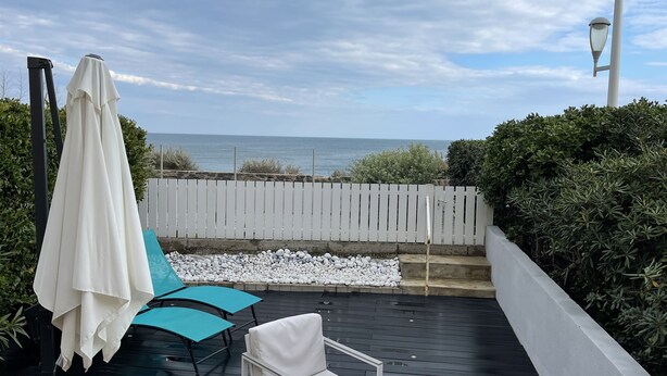 Une terrasse avec une chaise blanche, une chaise bleue et un parasol blanc. On y trouve une clôture blanche et une vue sur l’océan.