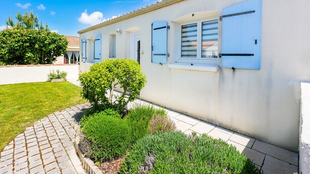 A single-story building with a white exterior and blue shutters, a small garden with a hedge and a tree, and a clear blue sky. - Charming 3-Bedroom House Near Beach with Garden and Terrace (Les Sables-d'Olonne)