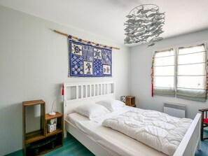 A bedroom with a white bed, a blue and white patterned tapestry, a fish sculpture, and a window with curtains.