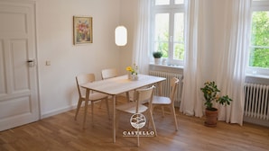 A dining area with a wooden table and chairs, a pendant light, a window with curtains, and a radiator.