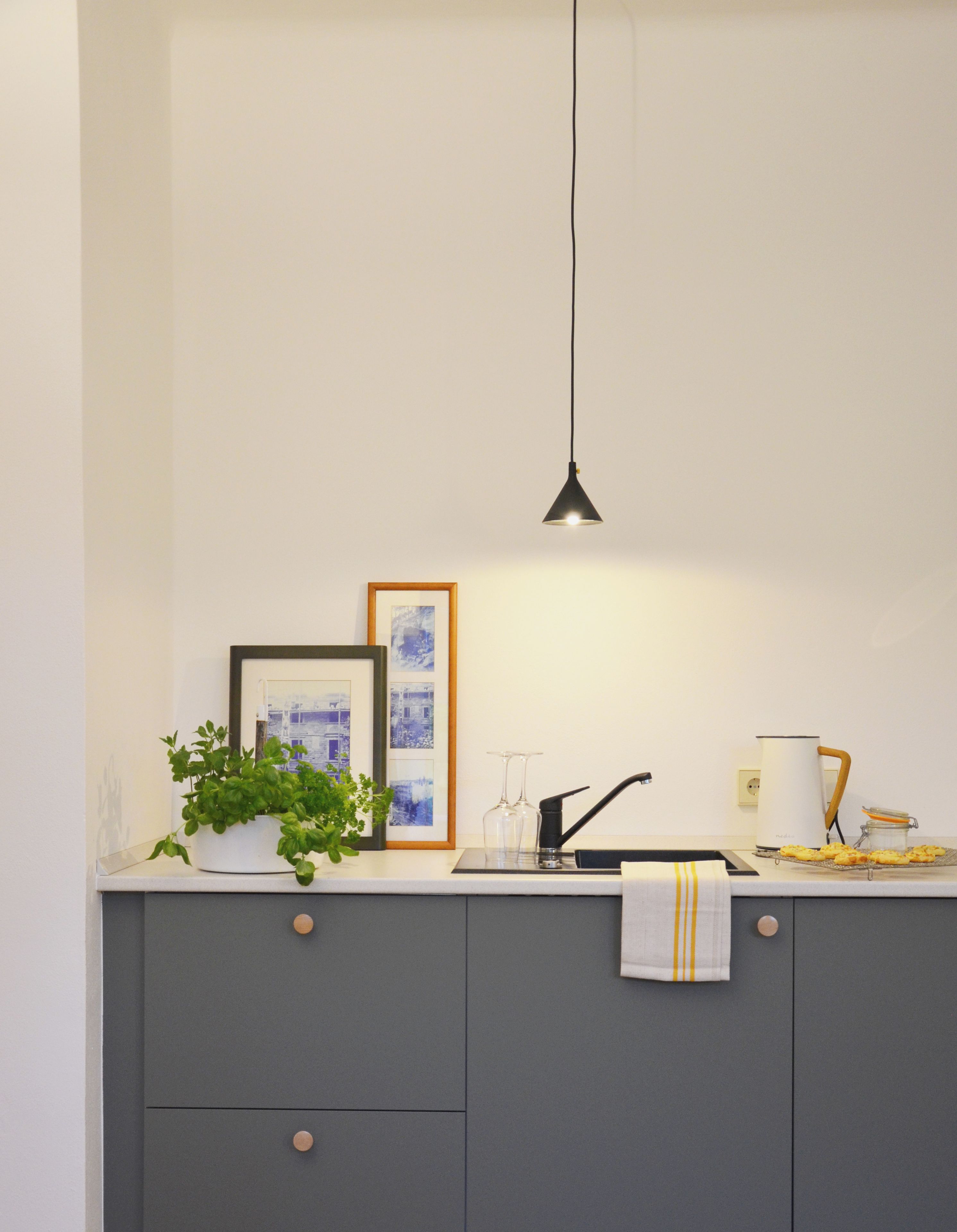 A modern kitchen with a grey cabinet, a hanging pendant light, a potted plant, framed pictures, and a towel on the counter.