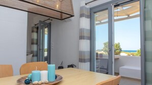 Een eetgedeelte met een tafel voor twee personen, uitzicht op de zee door een glazen deur, en een balkon met een witte bank.