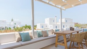 Een terras met een houten tafel en stoelen, een witte bank met kussens, en uitzicht op witte gebouwen op de achtergrond.