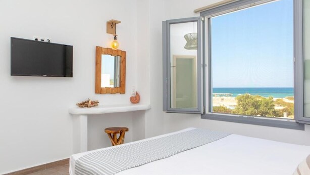 Een slaapkamer met een bed, een aan de muur bevestigde televisie, een klein houten krukje, een plank met decoratieve objecten en een raam met uitzicht op het strand.