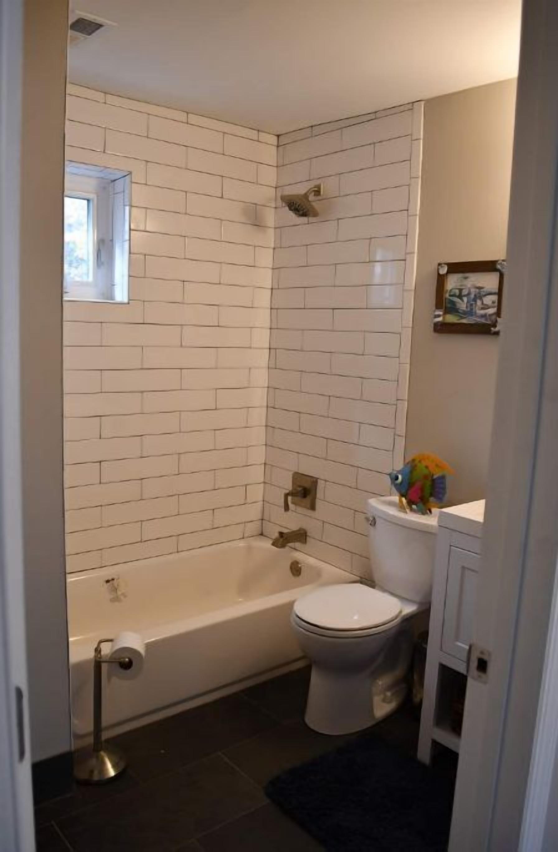Une salle de bain avec des carreaux de céramique blancs rectangulaires, des toilettes blanches, une baignoire blanche et une petite fenêtre située au-dessus des toilettes.
