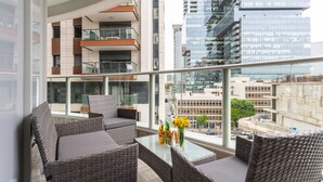 Ein Balkon mit Korbmöbeln, einem Glastisch und Blick auf eine Skyline mit modernen Gebäuden.