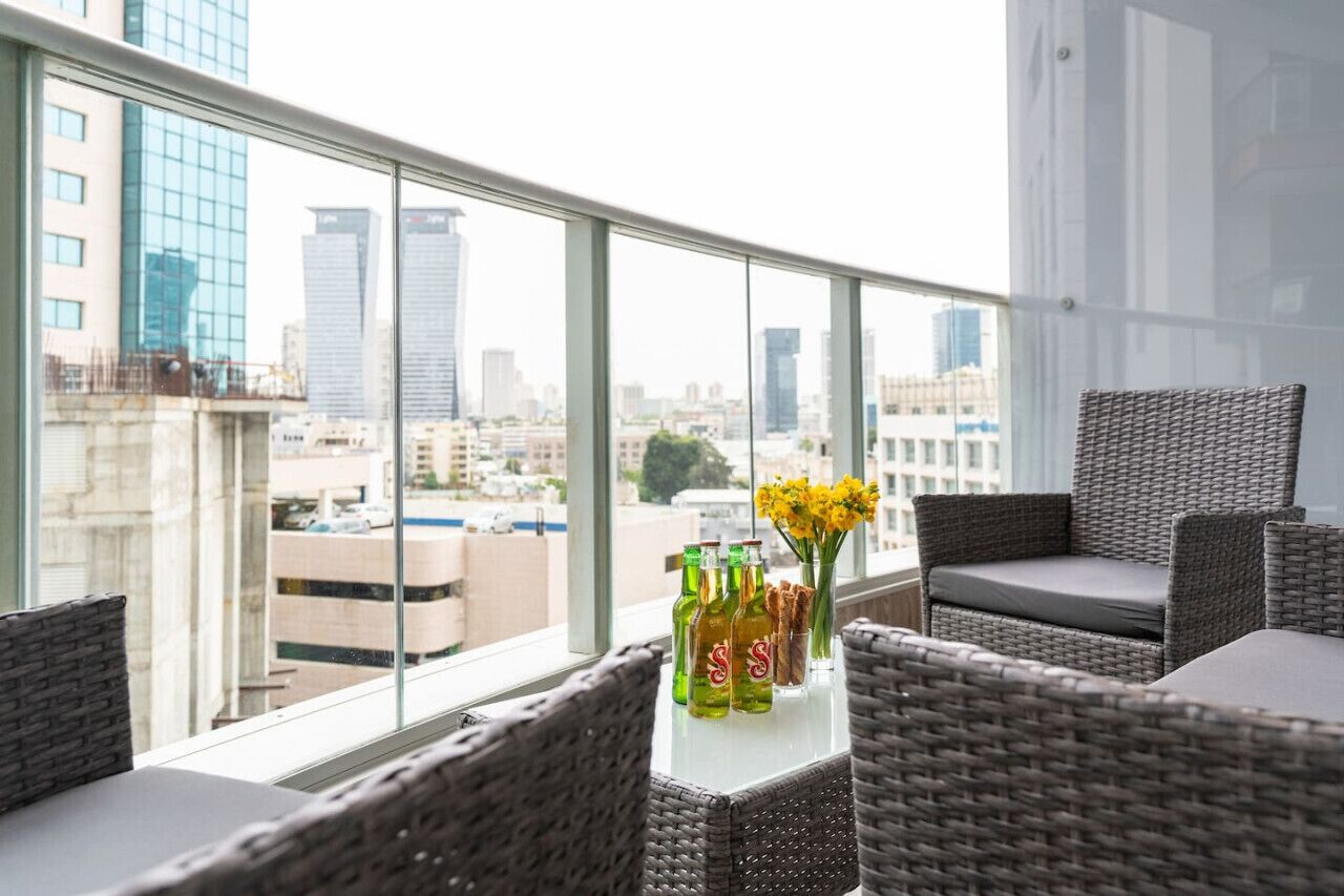 A balcony with wicker furniture, a table with bottles and flowers, and a city view through large windows.