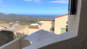 Terraço com vista para o mar e montanhas, um guarda-sol branco e um edifício com antena parabólica.