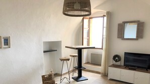 Uma sala de estar moderna com uma mesa branca, um banquinho trançado, uma TV sobre um móvel e uma luminária de palha no teto.