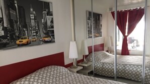 Une chambre d’hôtel avec un mur recouvert de miroirs qui reflète le paysage urbain, deux lits avec une literie à motifs, une table de chevet avec des lampes et une fenêtre avec des rideaux rouges.