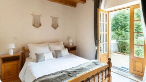 Ein Schlafzimmer mit einem hölzernen Bett, weißer Bettwäsche, zwei Nachttischen mit Lampen und Blick auf Grün hinter einem großen Fenster.