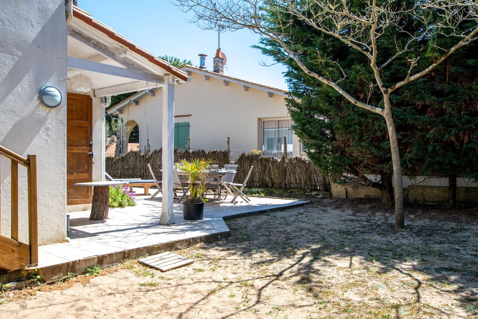 A house with a patio area, a wooden table and chairs, a potted plant, and a tree.