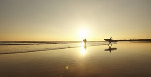 Une scène de plage au coucher du soleil, avec deux personnes qui se promènent et l’une d’entre elles porte une planche de surf.