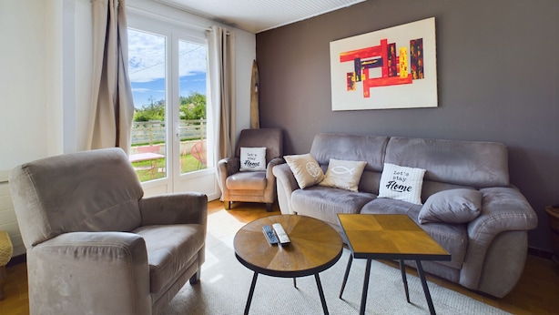 Ein Wohnzimmer mit grauem Sofa, grauem Sessel und einem kleinen Holztisch. An der Wand hängt ein Gemälde und das Fenster ist mit Vorhängen dekoriert.