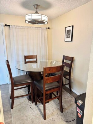 A dining area with a round glass table, four wooden chairs, a white curtain, a ceiling fan, and a framed picture on the wall. - Perfect Private Getaway in Seattle's Southside! (SeaTac)