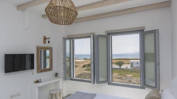 A bedroom with a bed, a TV, a mirror, a small table, and a window with a view of the beach. - Villa Lantana - Salinus Naxian Villas (Naxos)
