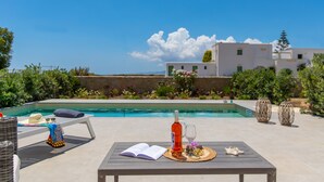A poolside area with a lounge chair, a table with a book, a bottle, and glasses, and a view of a white building with greenery. - Villa Lantana - Salinus Naxian Villas (Naxos)