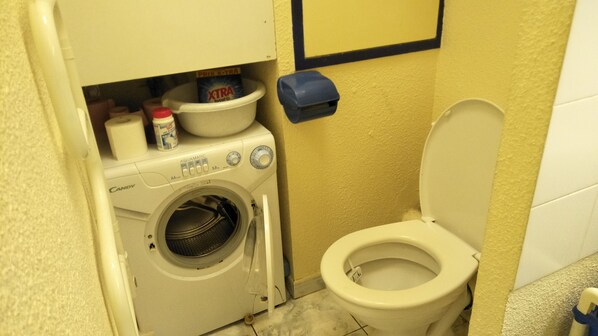 A compact bathroom with a washing machine, a toilet, and a trash bin.