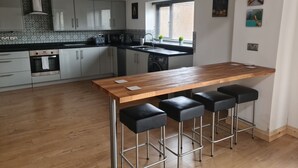 Een moderne keuken met een houten bar en zwarte barkrukken, witte kastjes en een raam met zonwering.