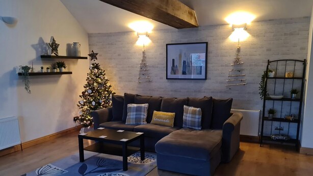 Een gezellige woonkamer met een donkergrijze bank, een salontafel en een kerstboom. Twee wandlampen, een afbeelding van een stadsgezicht in een lijst, en planken met planten en decoratieve objecten.