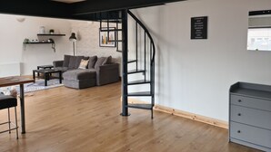 Een moderne woonkamer met een grijze bank, houten vloeren, een trap en een zwart wandplankje met planten.