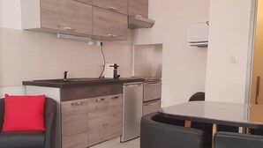 Une cuisine moderne avec des meubles en bois, un four encastré et un réfrigérateur compact. On y trouve également un coin repas avec une table et des chaises noires.