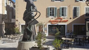 Une place pavée, ornée d’une sculpture, avec des tables et des chaises à l’extérieur, et un bâtiment dont la devanture est fermée par des volets.