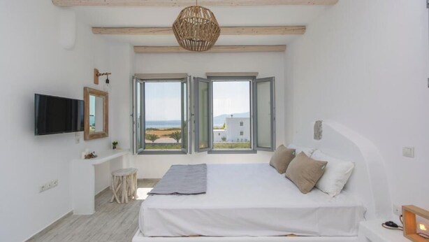Un dormitorio con cama, televisor, un taburete pequeño y una ventana con persiana abierta.