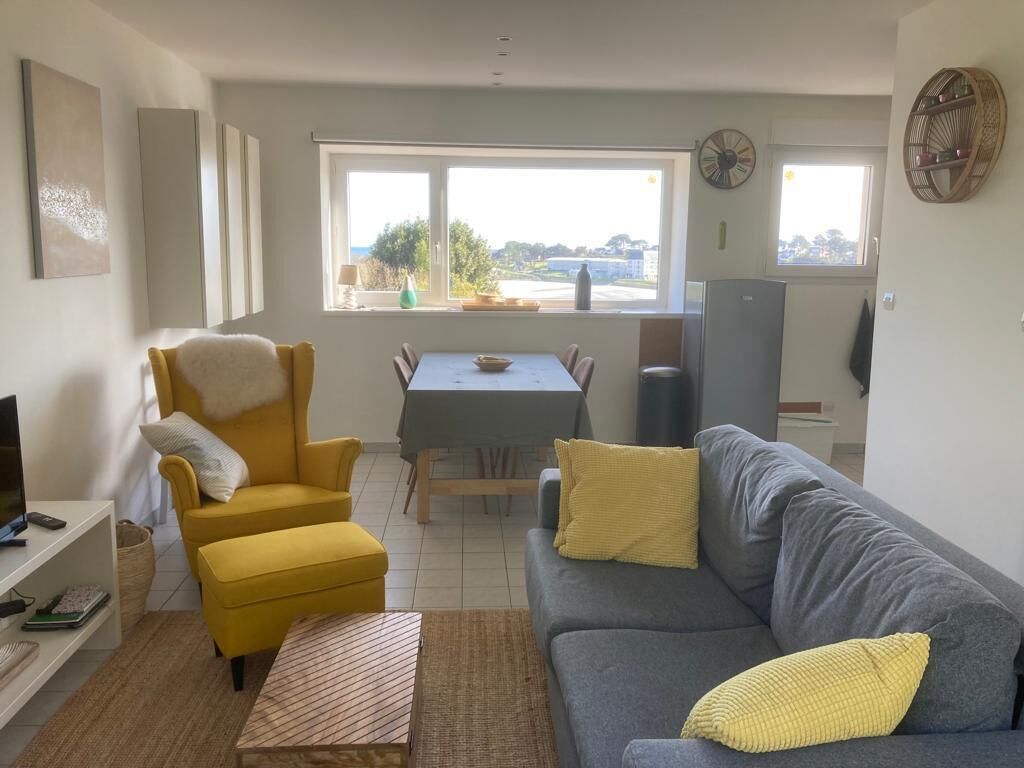 A living room with a sofa, armchair, and dining table. There is a window with a view of the outside.