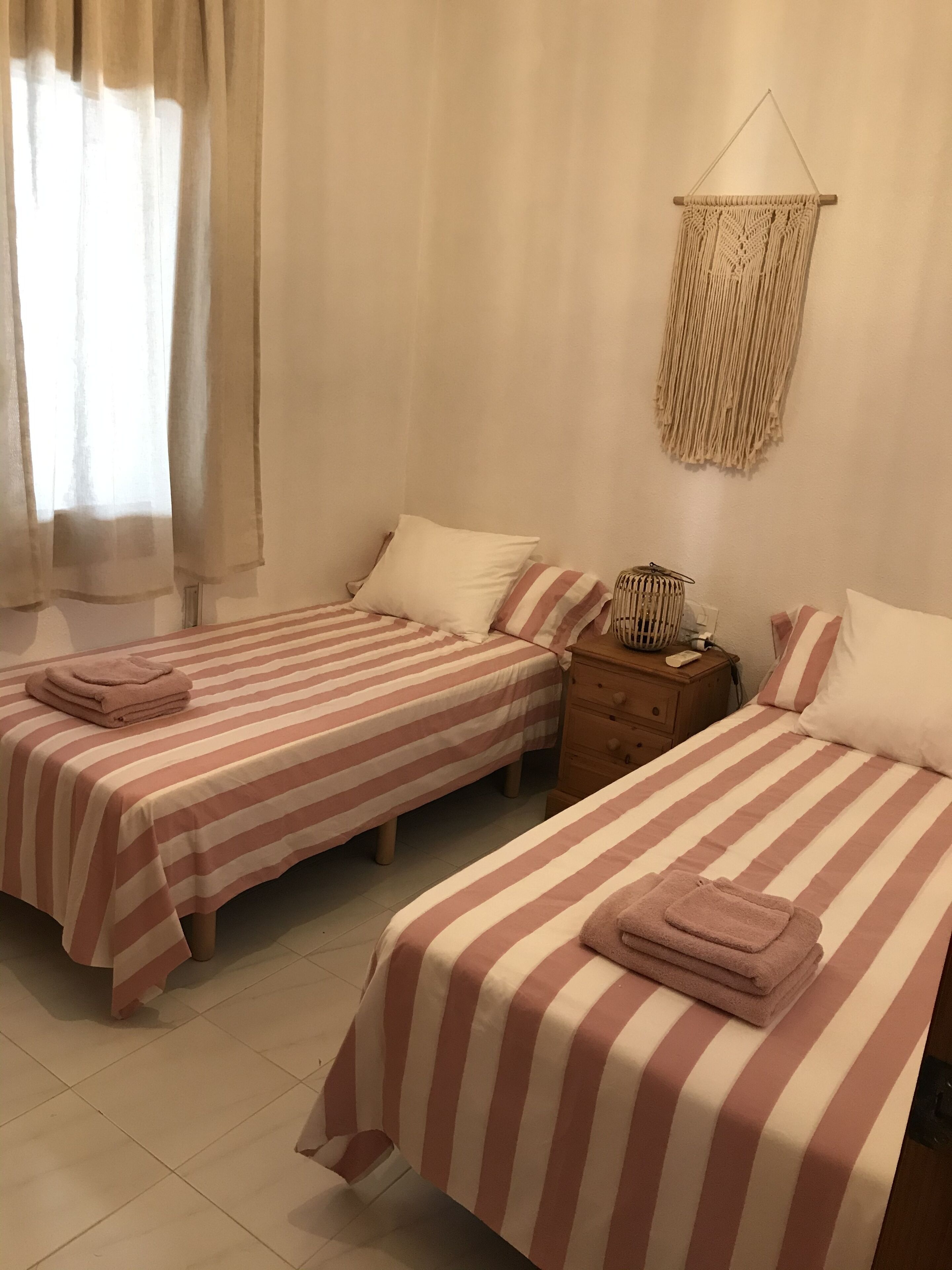 A room with two beds, each with striped bedding and pillows. There is a nightstand with a lamp and a macrame wall hanging.