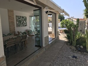 Un patio couvert avec des meubles en osier, une porte vitrée donnant sur une piscine et une vue sur les bâtiments voisins.