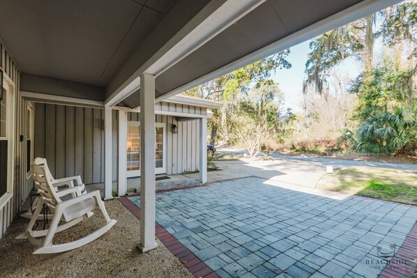 A covered patio with a rocking chair, a small building, and a paved walkway leading to a road.
