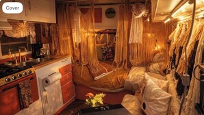 Een gezellige kamer met een bed, een klein tafeltje met bloemen, een keukenhoek en macramé decoraties.