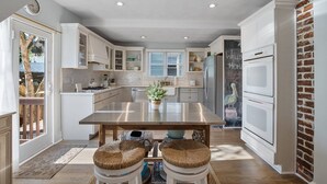 A modern kitchen with white cabinetry, a stainless steel island, and a brick accent wall. - Peach Perfect Victorian Home with Gourmet Kitchen (Cape Charles)