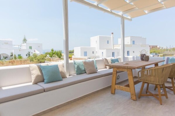 Una terraza con mesa y sillas de madera, un sofá blanco con cojines y vistas a edificios blancos al fondo.