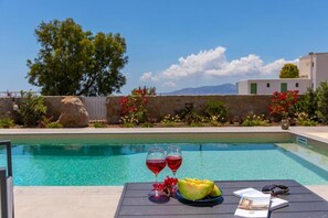 Escena junto a la piscina con una mesa, dos copas de vino tinto, un plato de melón y gafas de sol.