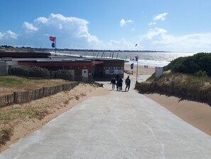 A beachside walkway leading to a building with a red roof, a sandy path, and a view of the sea.