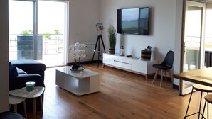 Ein geräumiges Wohnzimmer mit großem Fernseher, Holzboden, einer Couch, einem Couchtisch und einem Essbereich mit Stühlen.