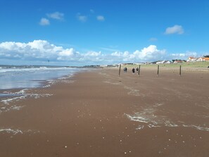 A wide sandy beach with gentle waves, a clear sky, and a few people walking.