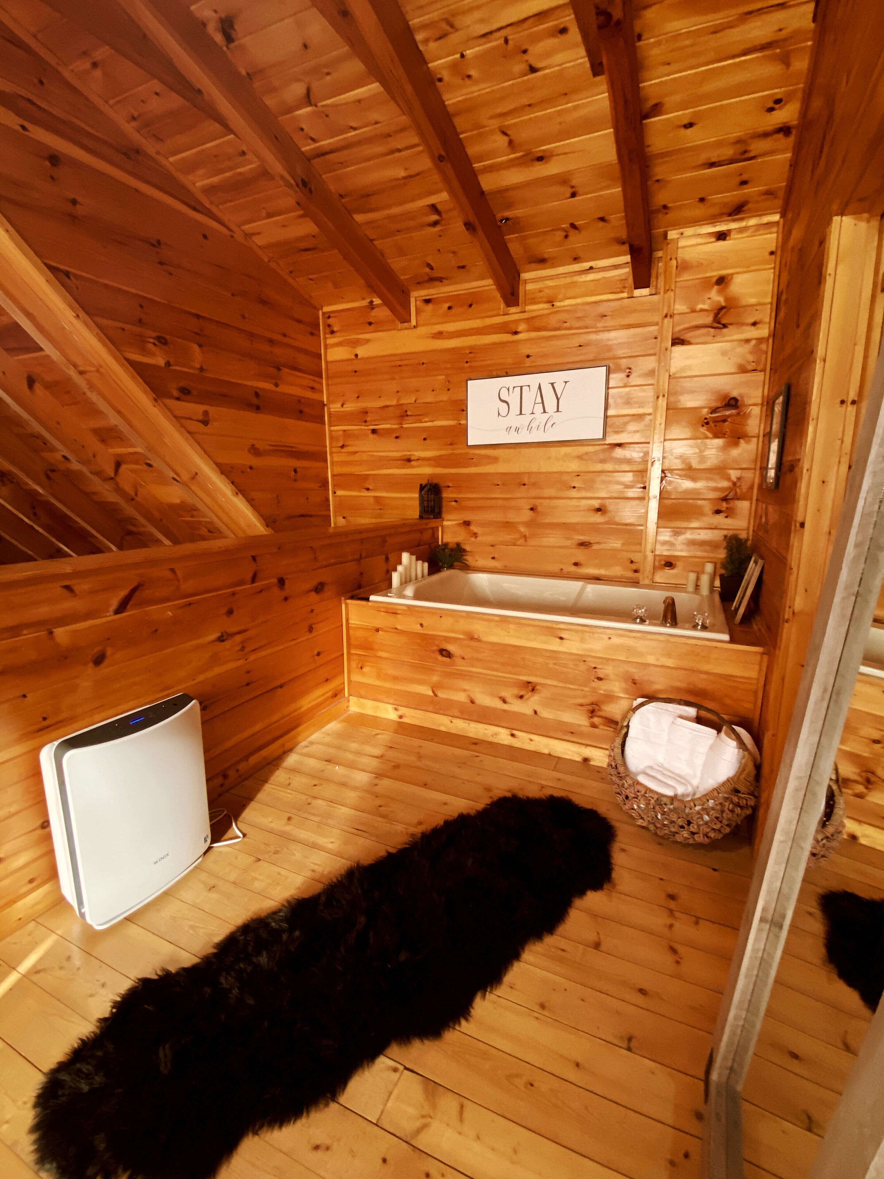 A wooden cabin interior with a wooden ceiling, a sign that says 'STAY an' Bilo', a bathtub, a white portable heater, and a basket with a white towel.