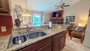 A kitchen with granite countertops, stainless steel sink, and wooden cabinets. There is a ceiling fan, a TV, and a painting on the wall.