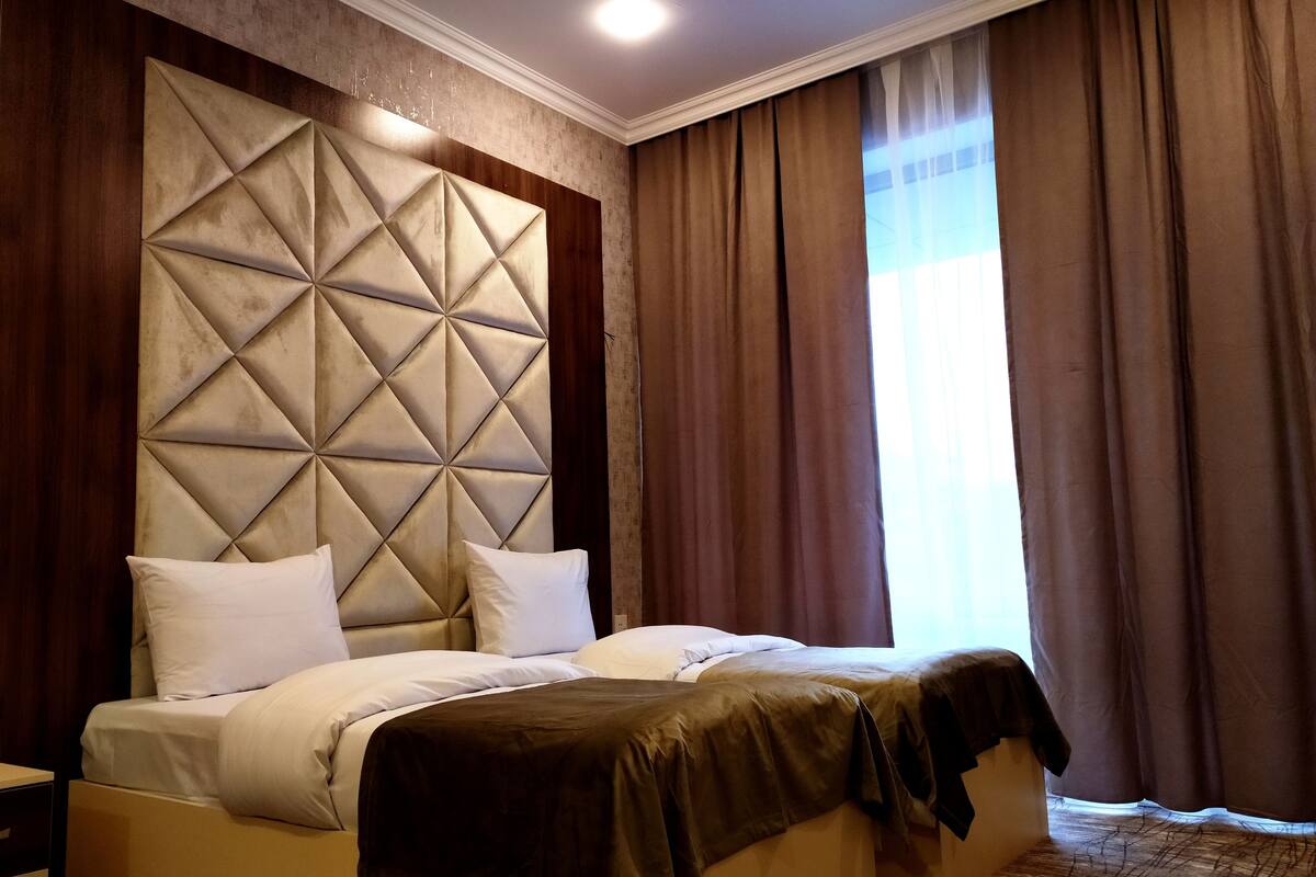 A hotel room with a large bed, a padded headboard, and a view of the outdoors through curtains.