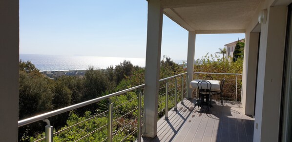 Un balcón con mesa y sillas, con vista a un paisaje costero con árboles y el mar.
